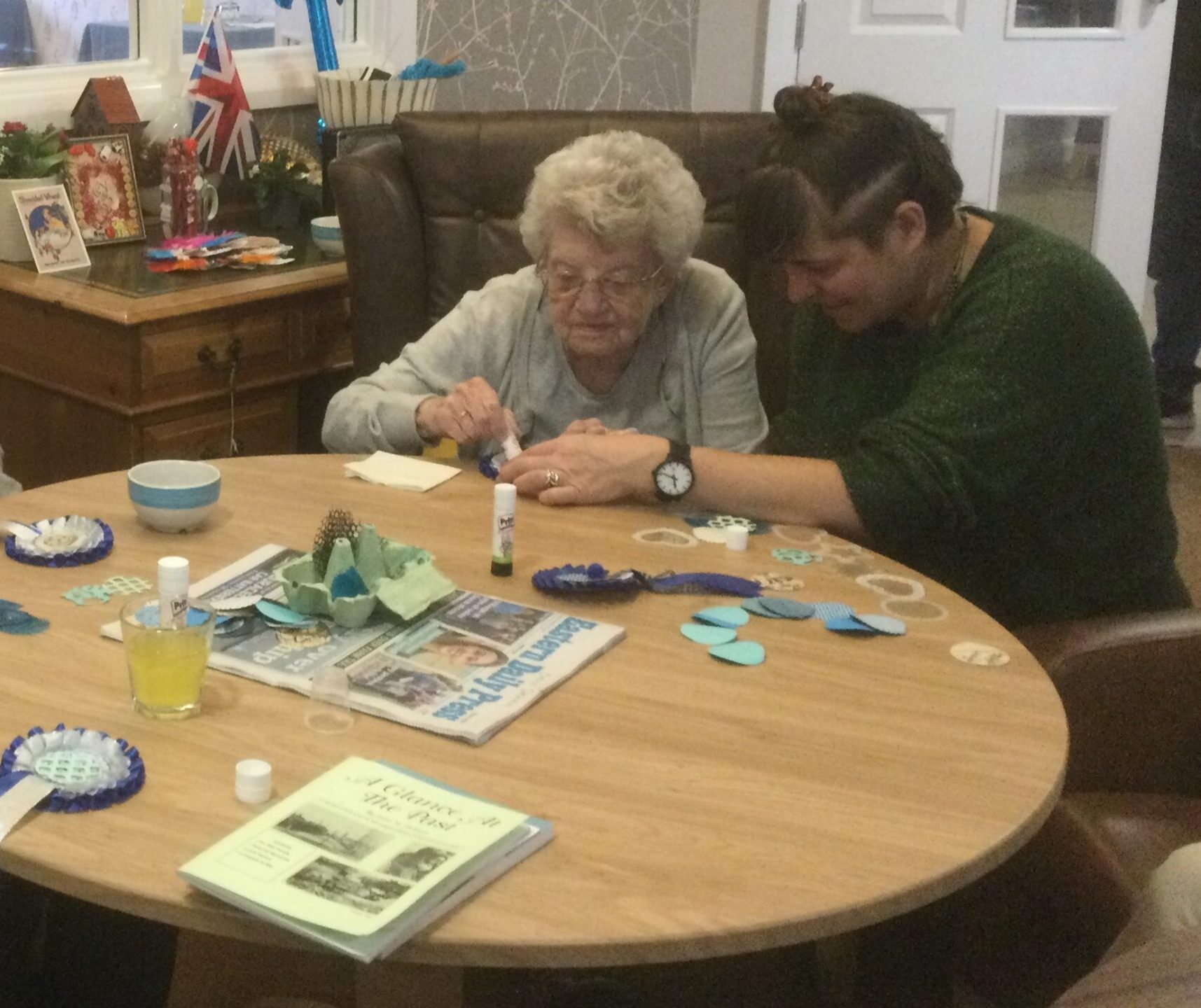 Two women seated side by side by a round table covered in craft materials. One woman steadies a piece of work, whilst the other woman glues something into place