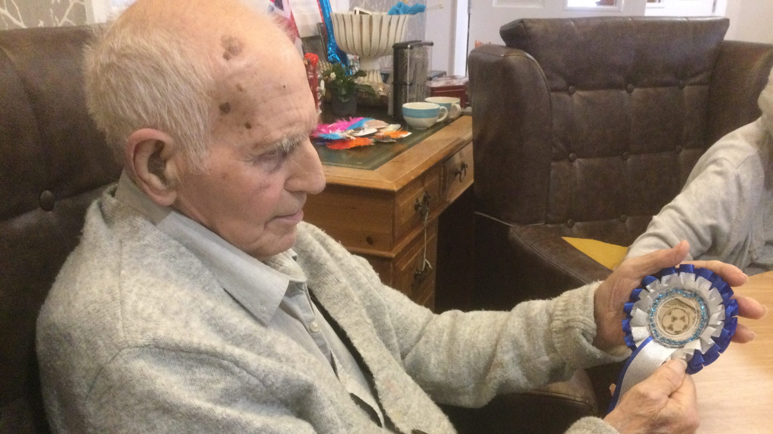 An elderly gentleman, seated, holding a blue and white rosette with a central image of a football