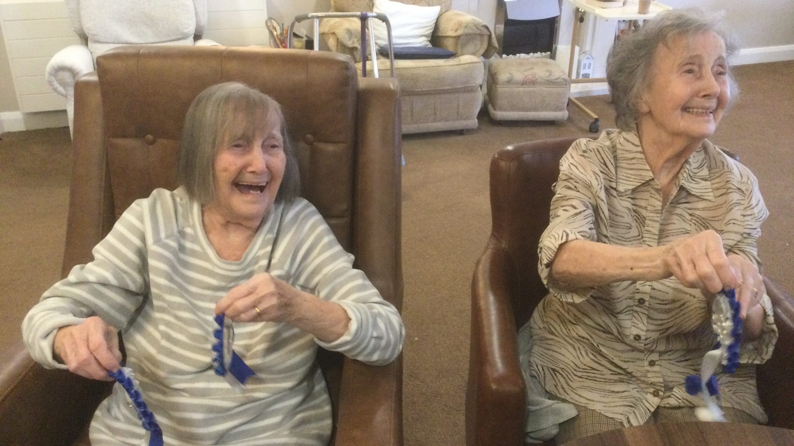 Two women seated side by side in brown chairs, holding rosettes and laughing