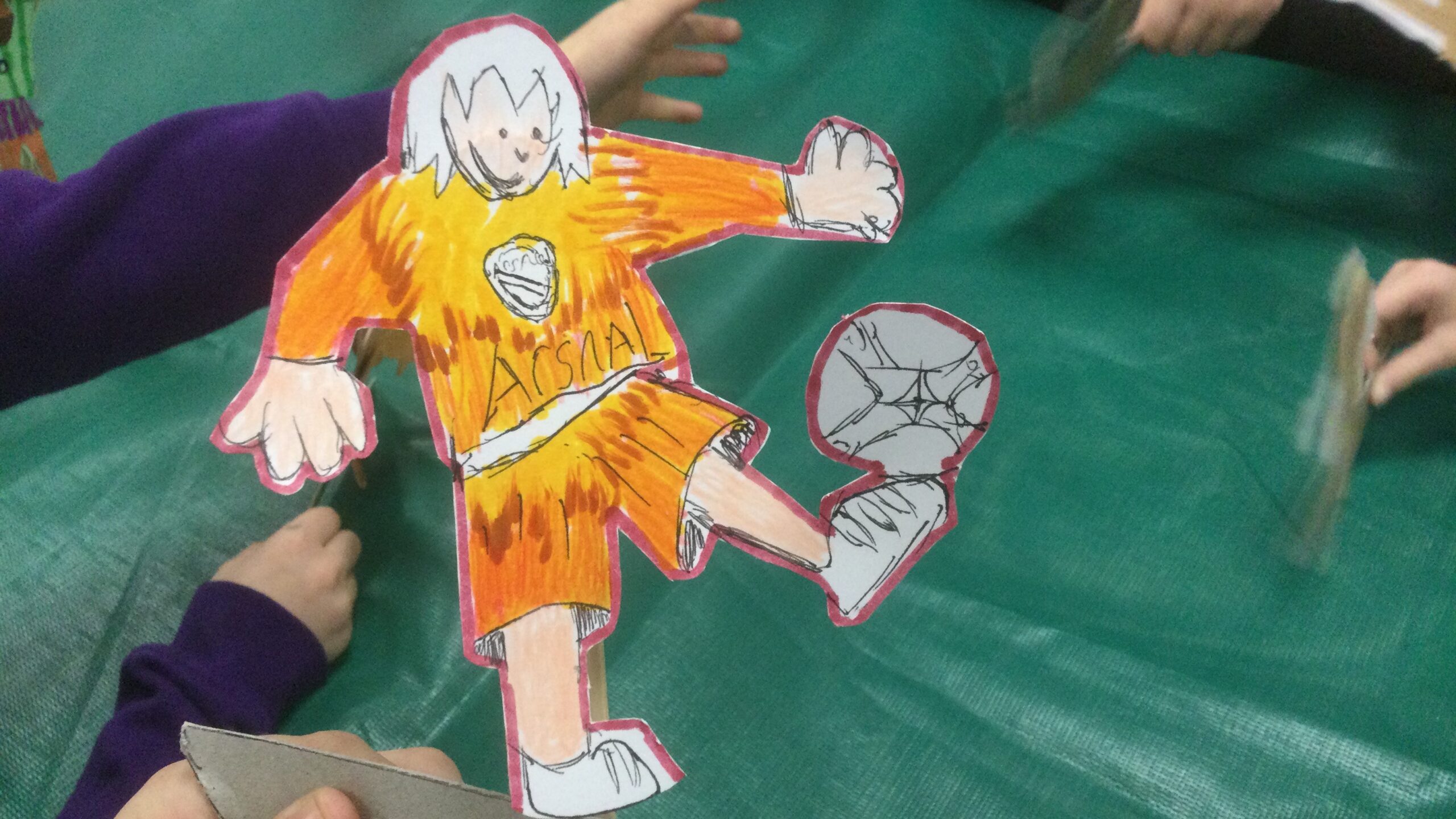 A flame coloured football figure balancing a ball on his toe. There are hands in the background