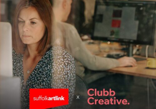 A woman and a man are working on computers with large monitors in an office. The woman is facing towards the camera and the man is facing away. The photo includes Suffolk Artlink and Clubb Creative's logos.