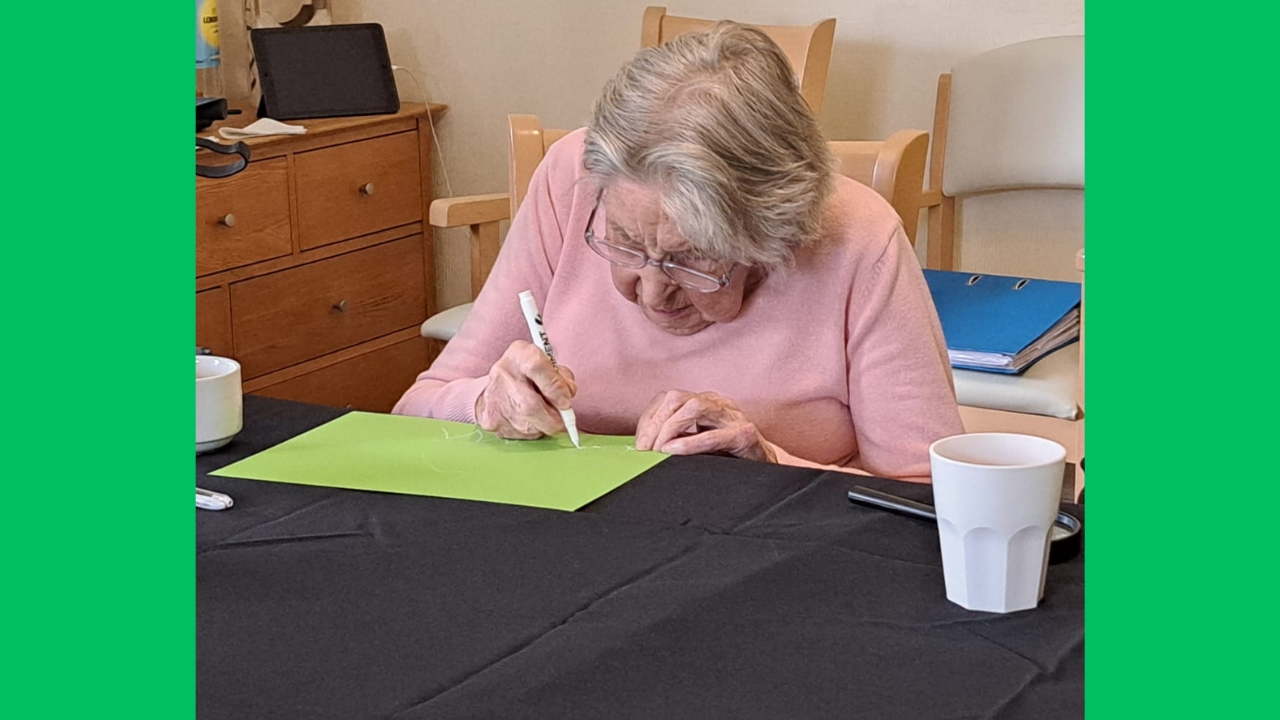 A woman in a pink jumper leans over a green piece of paper and draws on it in white pen