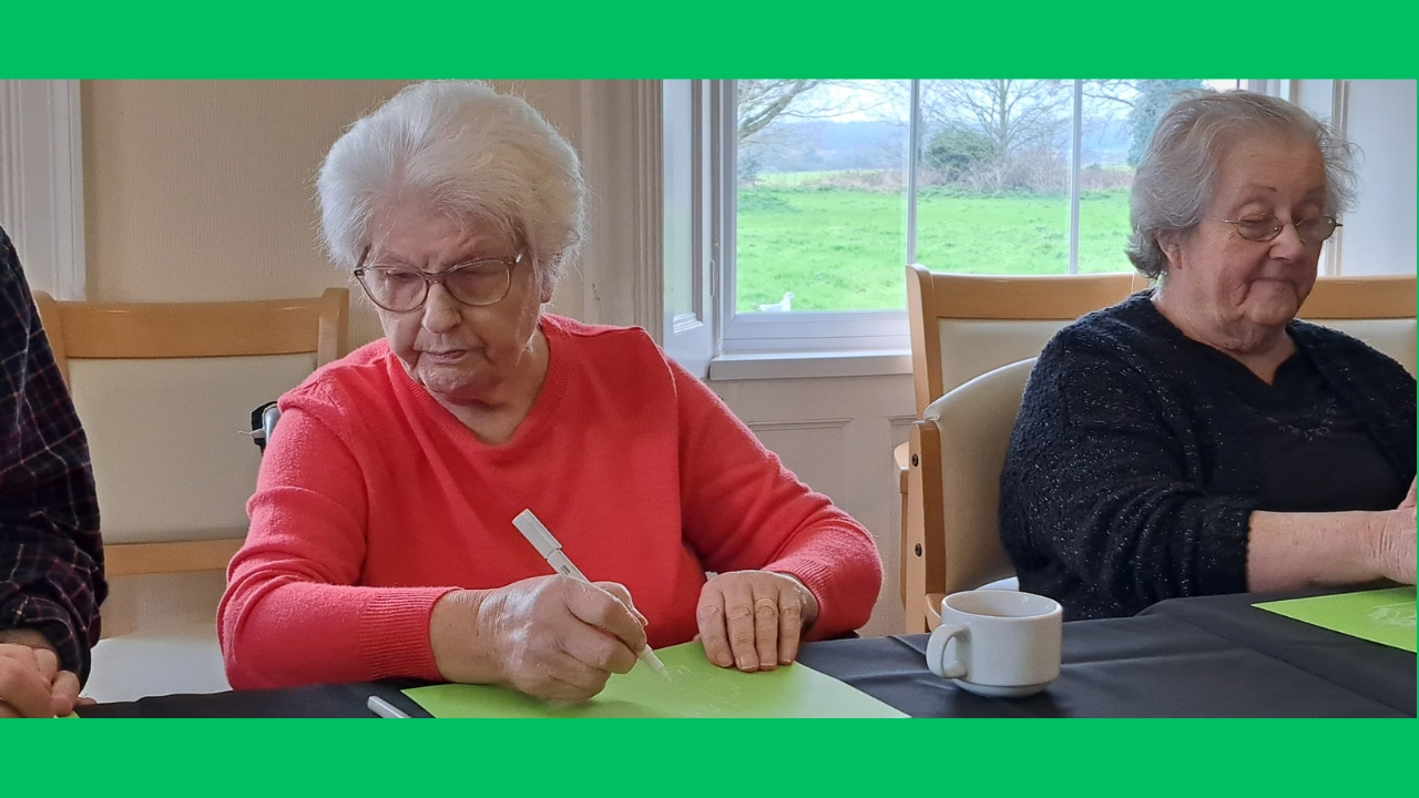 A woman in a bright orange jumper seated next to a lady in a dark top, each with a sheet of green paper in front of them and drawing with white pens. A window behind has a view across a large garden with fields beyond