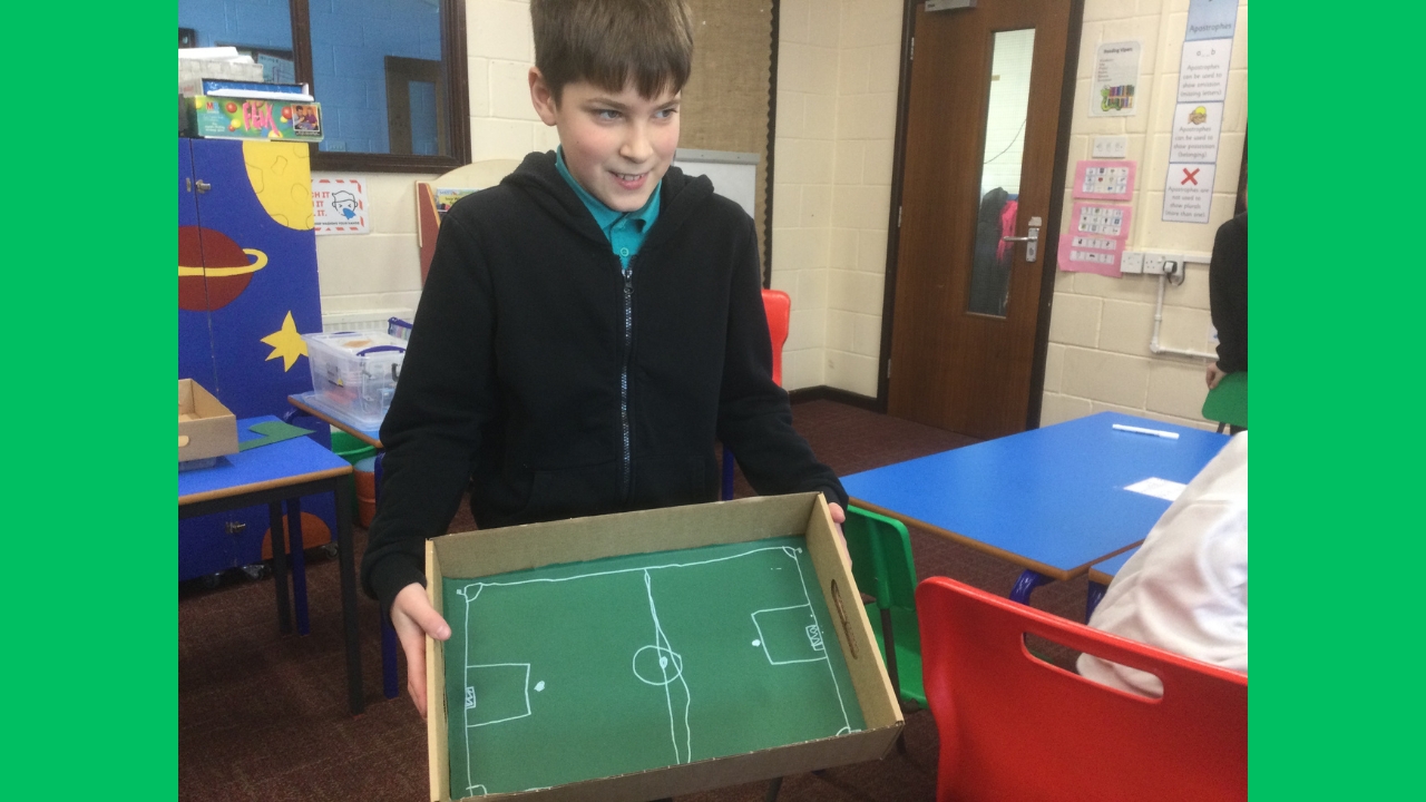 A school boy wearing a dark zipped jersey with green T-shirt beneath holds a shallow brown cardboard box with green inlay and drawn up to represent a football pitch