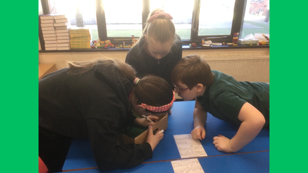 Three school children engrossed in drawing something inside a shallow cardboard box. There is a window behind, with stacks of books, Lego bricks and other objects on the sill