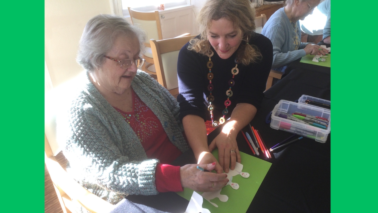 A woman with hair in bunches and wearing a colourful long loop necklace holds a row of paper people steady whilst another woman adds features to their faces