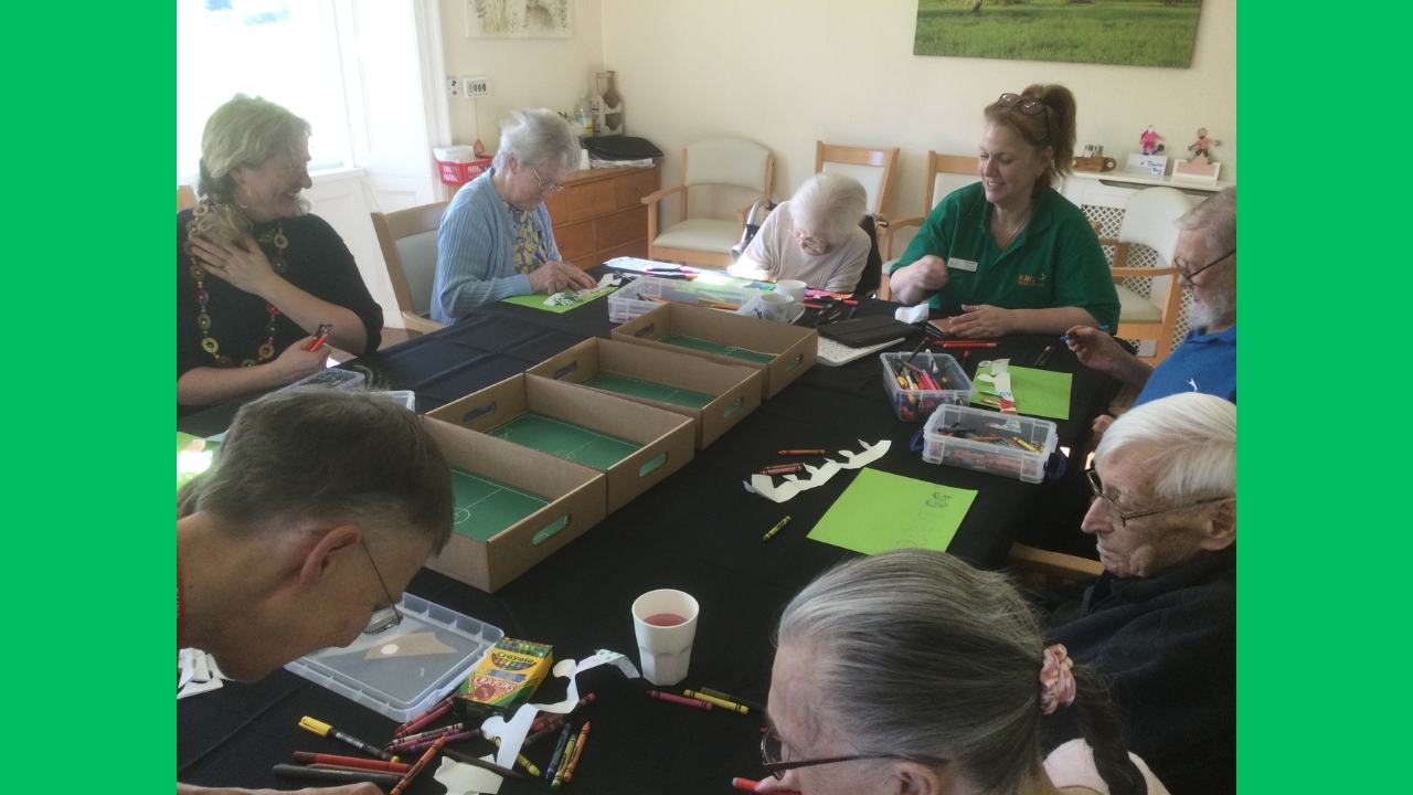 A group of adults around a table covered in 4 shallow cardboard boxes, each with a green football pitch interior, plus pens drinks and sheets of green paper