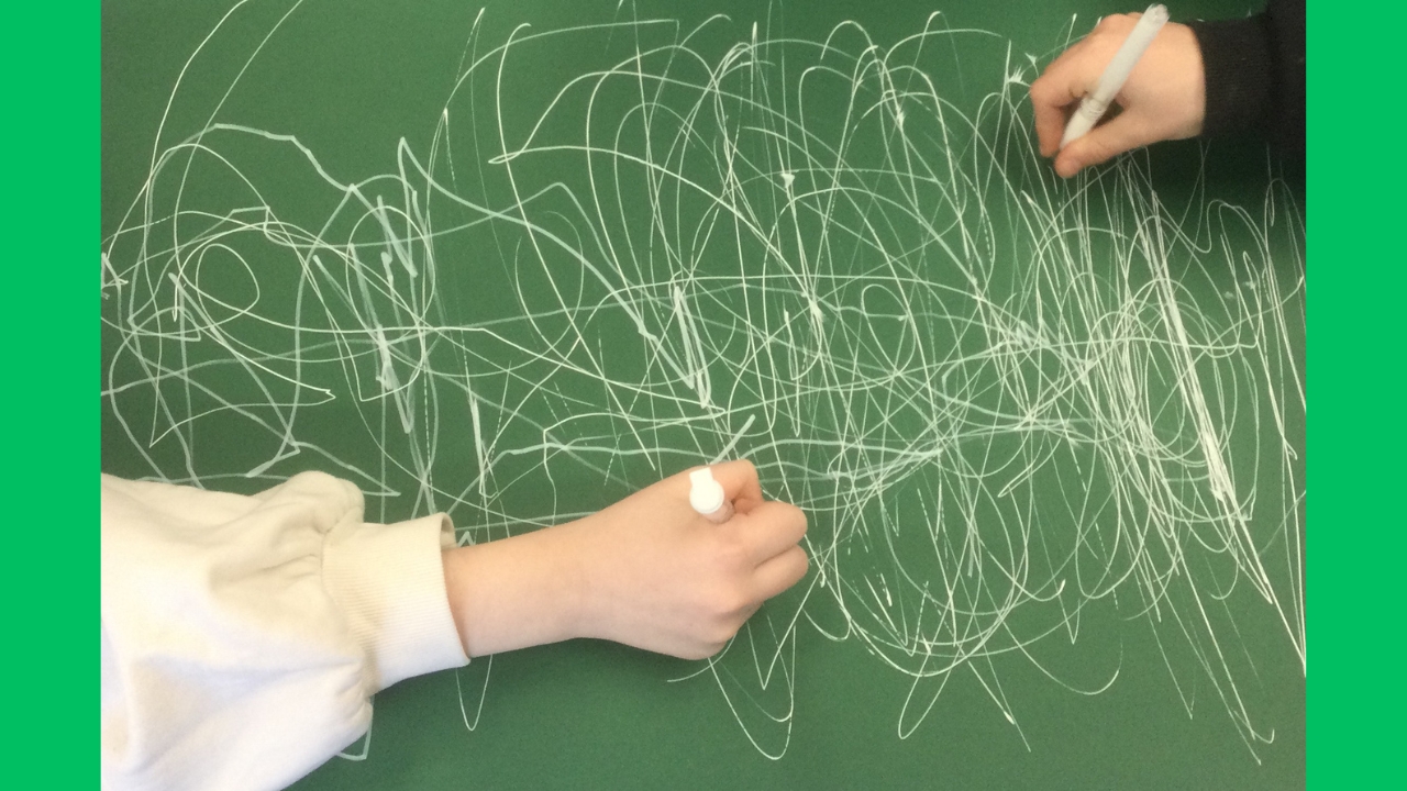 Overhead view showing hands at opposing ends of a large sheet of green paper, each holding a white pen and lots of white lines across the page