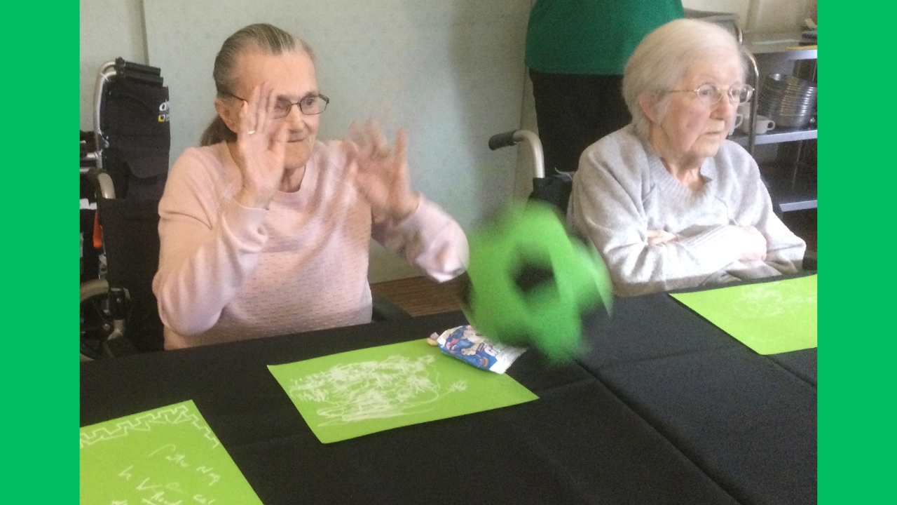 A woman has her hands up in the air, having just launched a black and green foam football across the table. Her companion is seated beside her, looking across the table