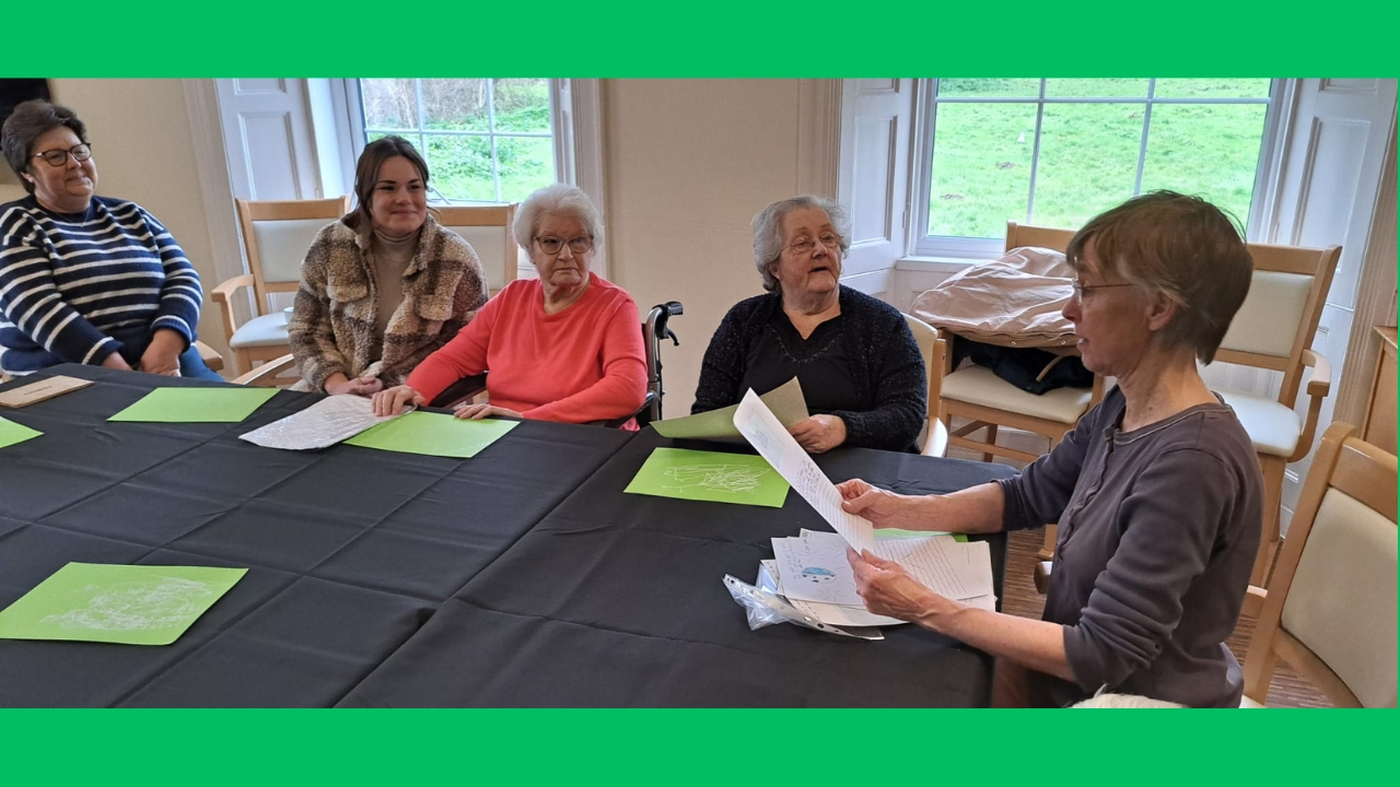 A group of five women around a table, looking at letters and pictures. Two large windows behind look out over a large green lawn.