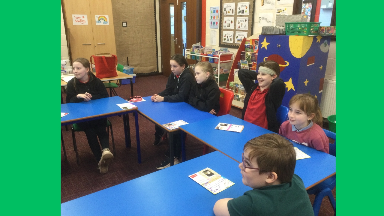 A group of 6 school children seated at blue tables clearly listening to someone, as they are focused in one direction and smiling.