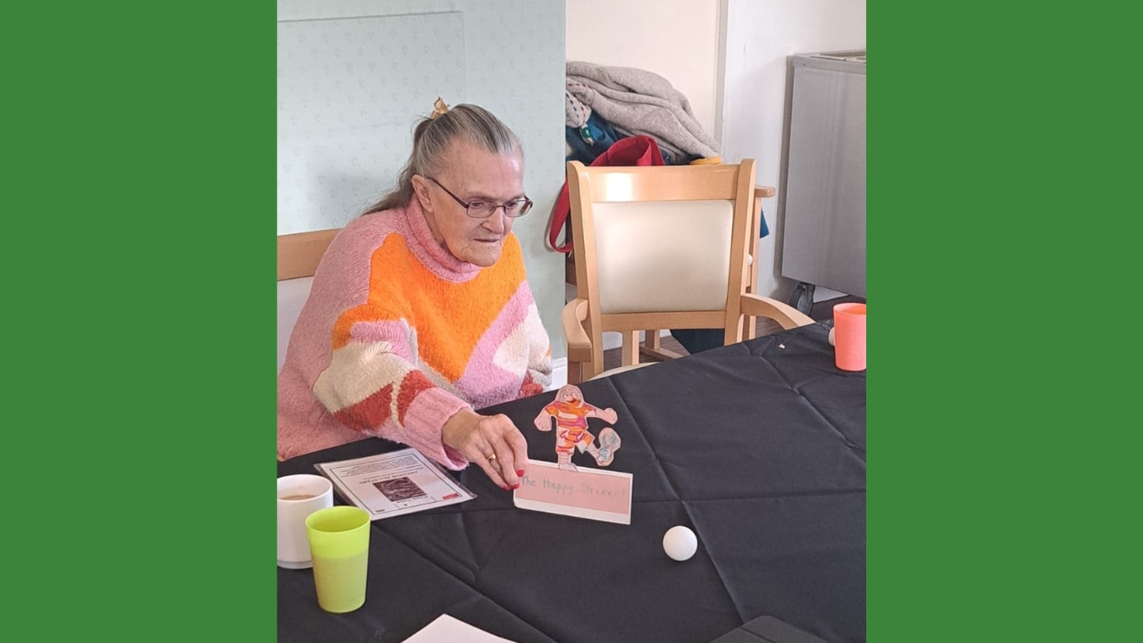 A woman in a colourful pink and orange jumper has made a football character in the same colours and is using it to flick a ping pong ball across a table covered in a black cloth