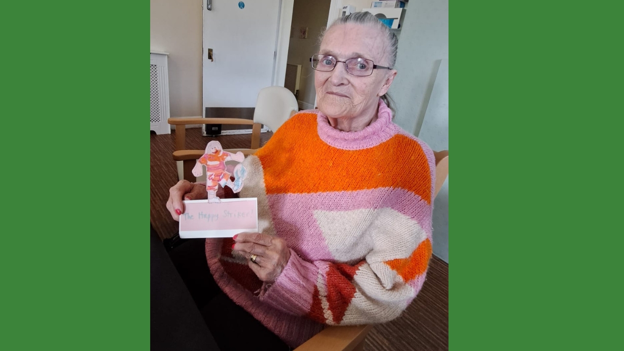 A woman in a colourful pink and orange jumper holds u[ a cardboard football character in similar colours