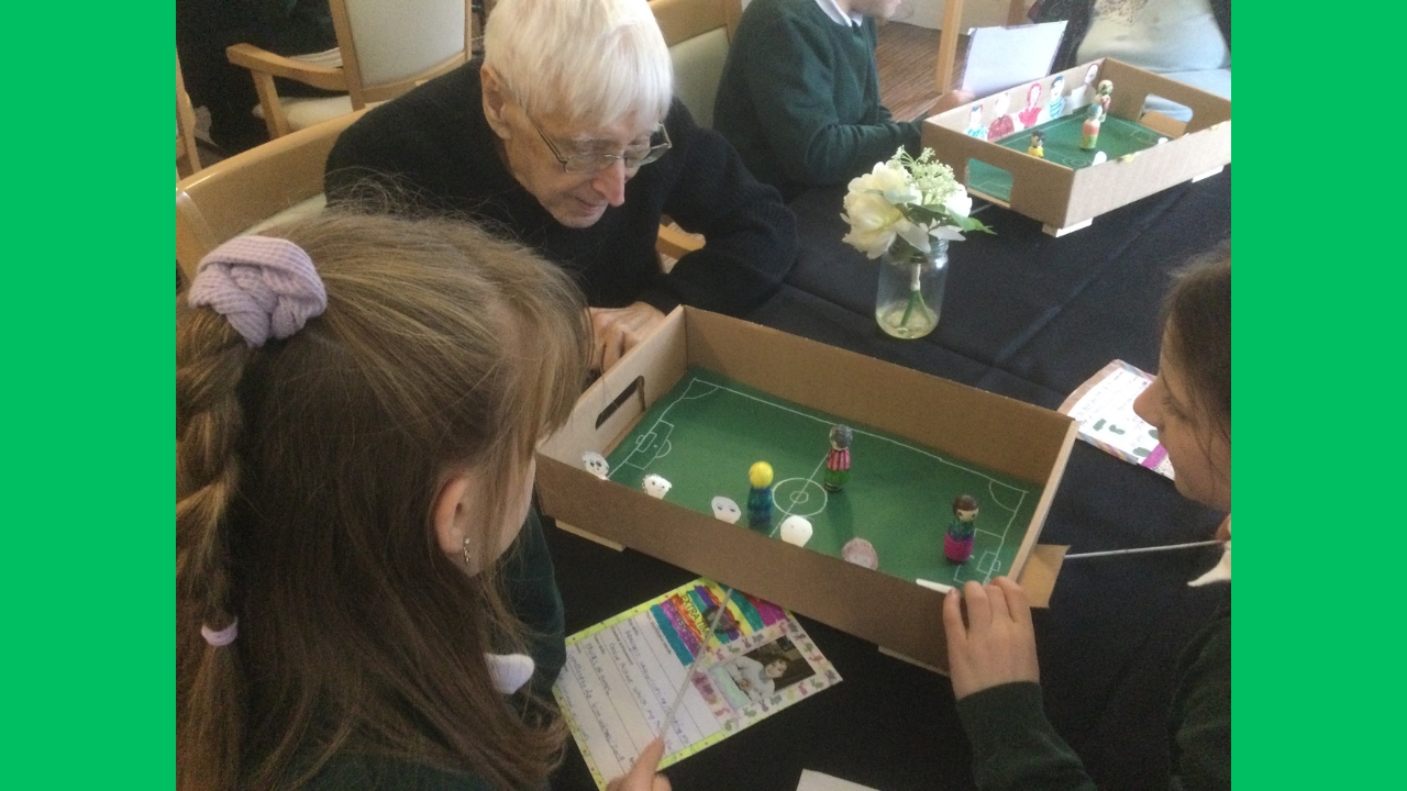 A gentleman looks over a cardboard box football pitch on which are three small wooden figures. Two school girls sit beside him and there's a vase of white roses to his left