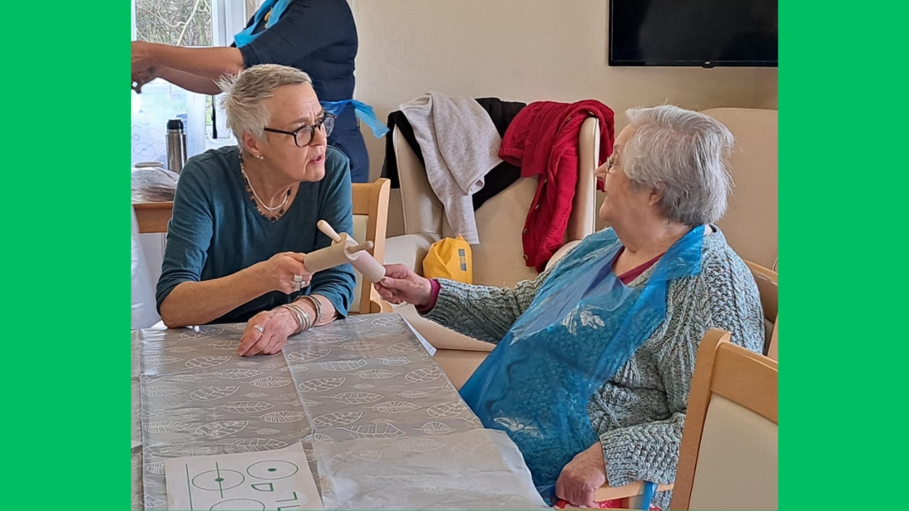 Two women seated facing each other at a table, one wearing a blue plastic apron. They are holding small rolling pins and have them crossed in front of them