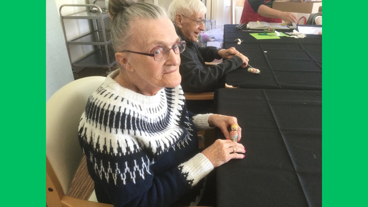 A woman wearing a blue and white pattern knitted jumper is seated at a table covered in a black cloth - she is holding a small wooden figure