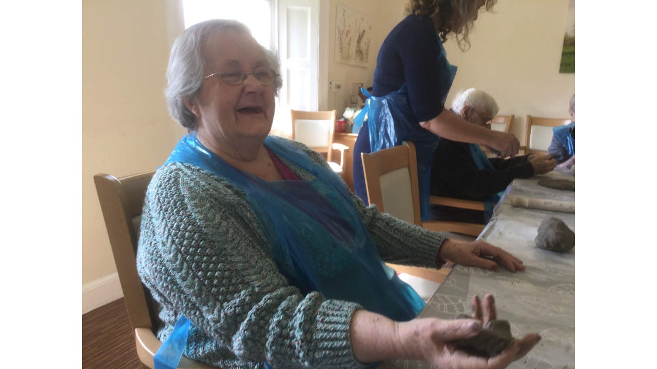 A woman smiles at the camera. In her right hand is a small ball of clay whilst another is on the table to her left and at the other end of the table are more people using clay