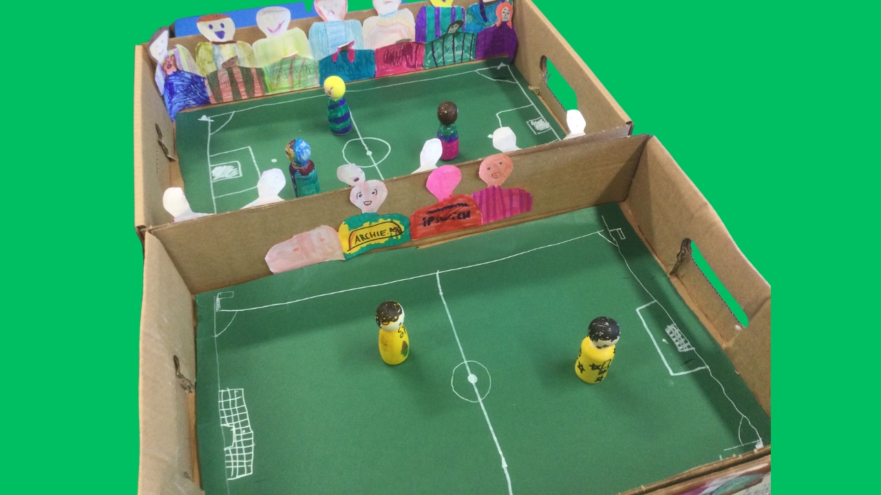 Two cardboard football pitches with brightly coloured wooden figures on each and hand drawn supporters glued to the sides