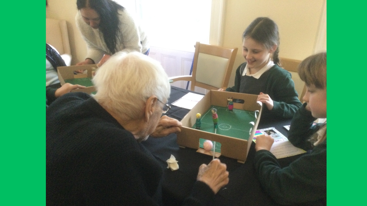 A child grins as she plays football, a second child looks on and a gentleman attempts to block the goal mouth of the magnetic football game