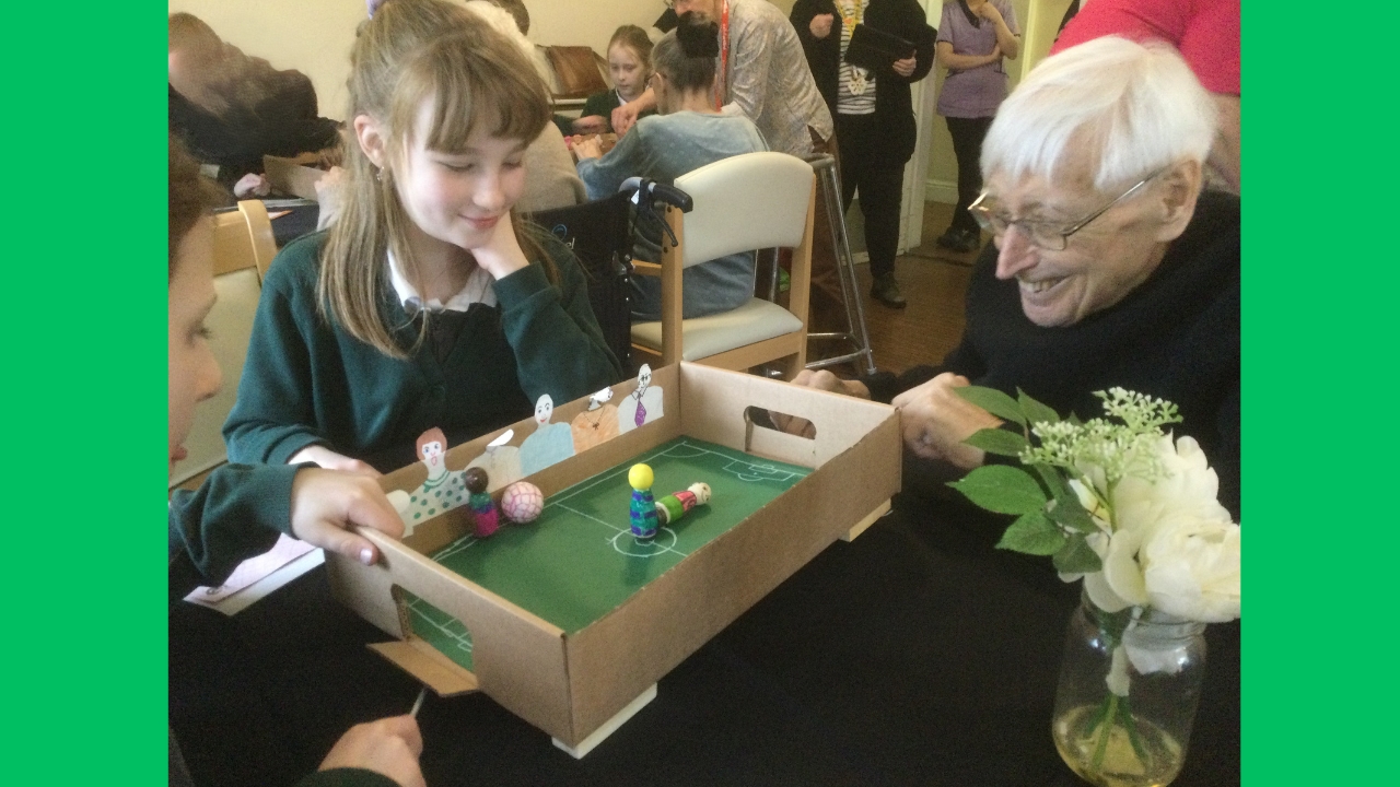 A gentleman grins broadly as he plays a game of magnetic football with two school girls. There are white flowers in the foreground and behind are several people, some seated, some standing