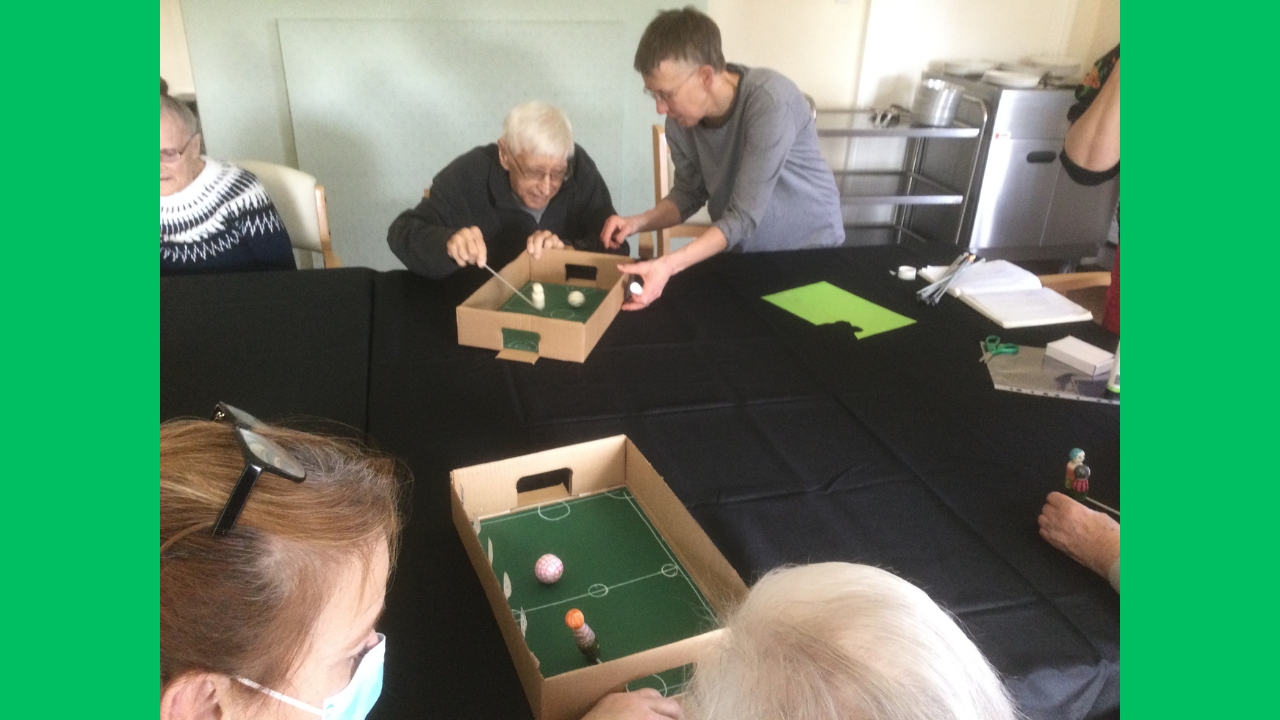 A woman steadies a cardboard box whilst a gentleman manoeuvres a figure on a stick across the surface