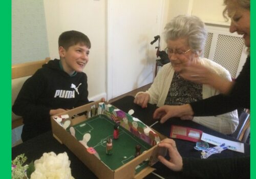 A boy laughs as two women, one seated, one standing attend to a magnetic football game on the table in front of them