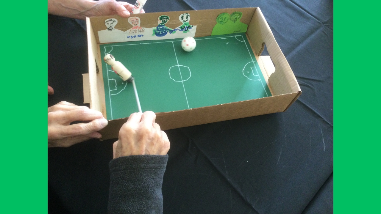 Closs up of a cardboard football pitch with paper supporters glued to the sides and a magnetic player kicking the ball towards the goal mouth