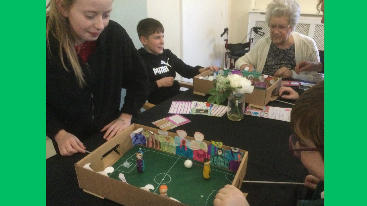 Children seated and standing at a table with two hand made magnetic football games in front of them on a black cloth. A lady sits one end, focused on the game