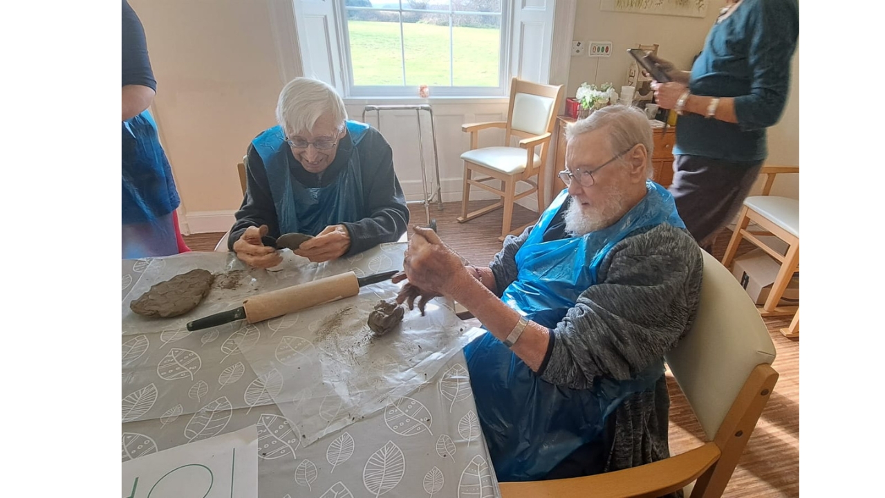 Two gentlemen, each wearing a blue plastic apron are softening lumps of clay in their hands whilst on the table is a rolling pin and some plastic mats