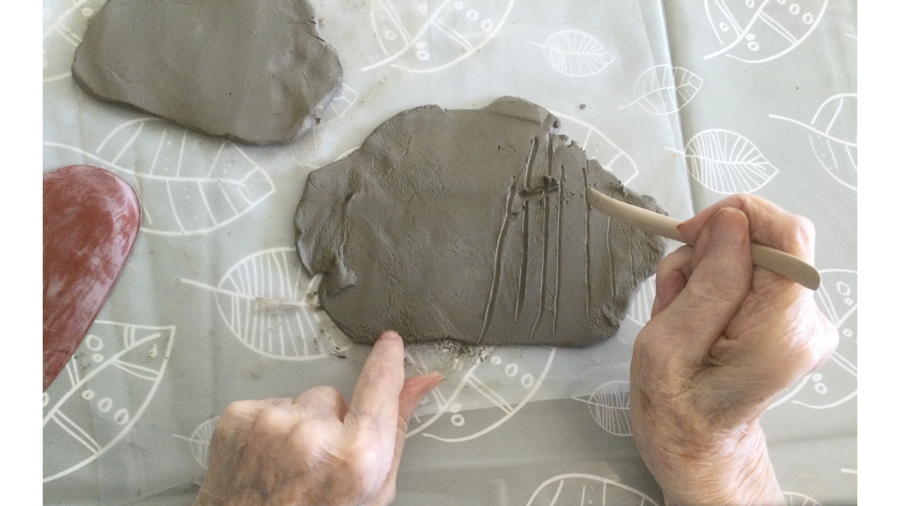 Overhead view of a pair of hands with two greyish flattened pieces of clay in front and a red 'kidney', a tool for smoothing clay, to the left. In the right hand there's a wooden implement which is being used to pull vertical lines through the clay