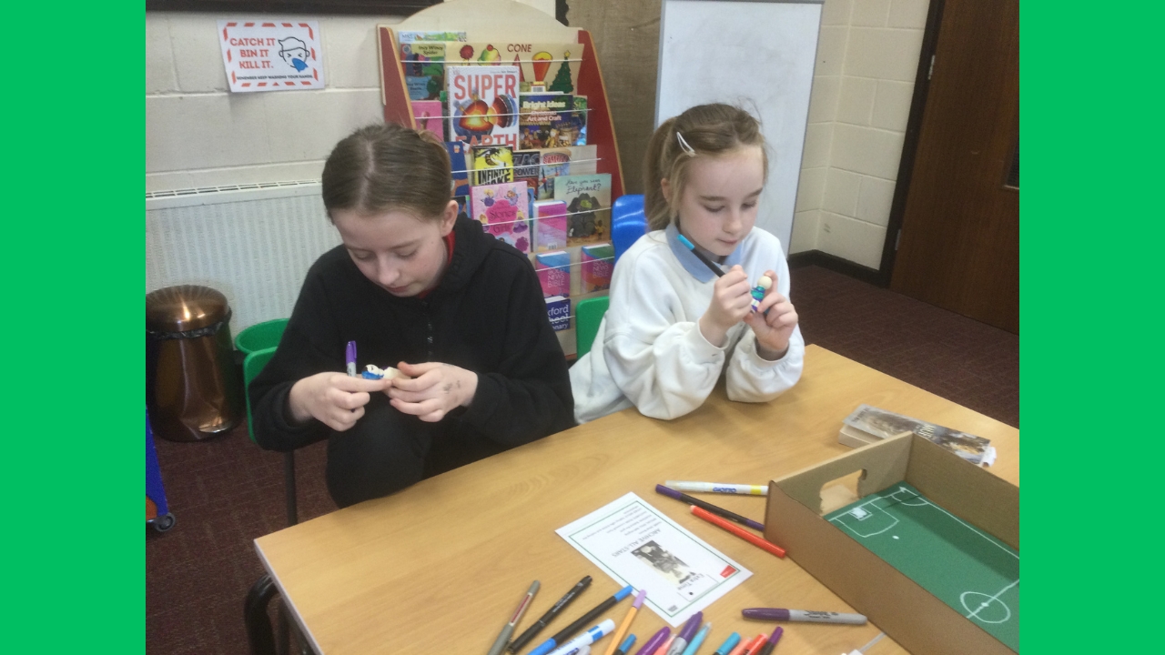 Two school girls seated at a table focused on colouring their wooden figures. In the background is a book case