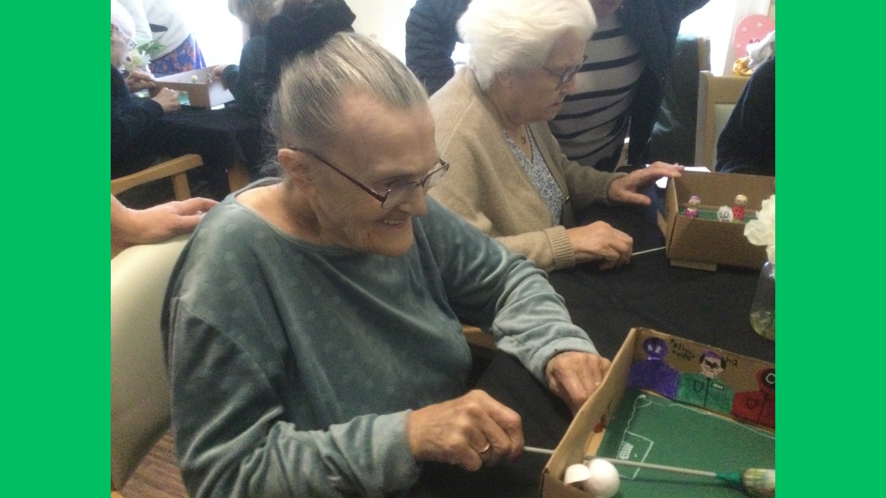 Two women seated side by side playing table top magnetic football, one grinning broadly