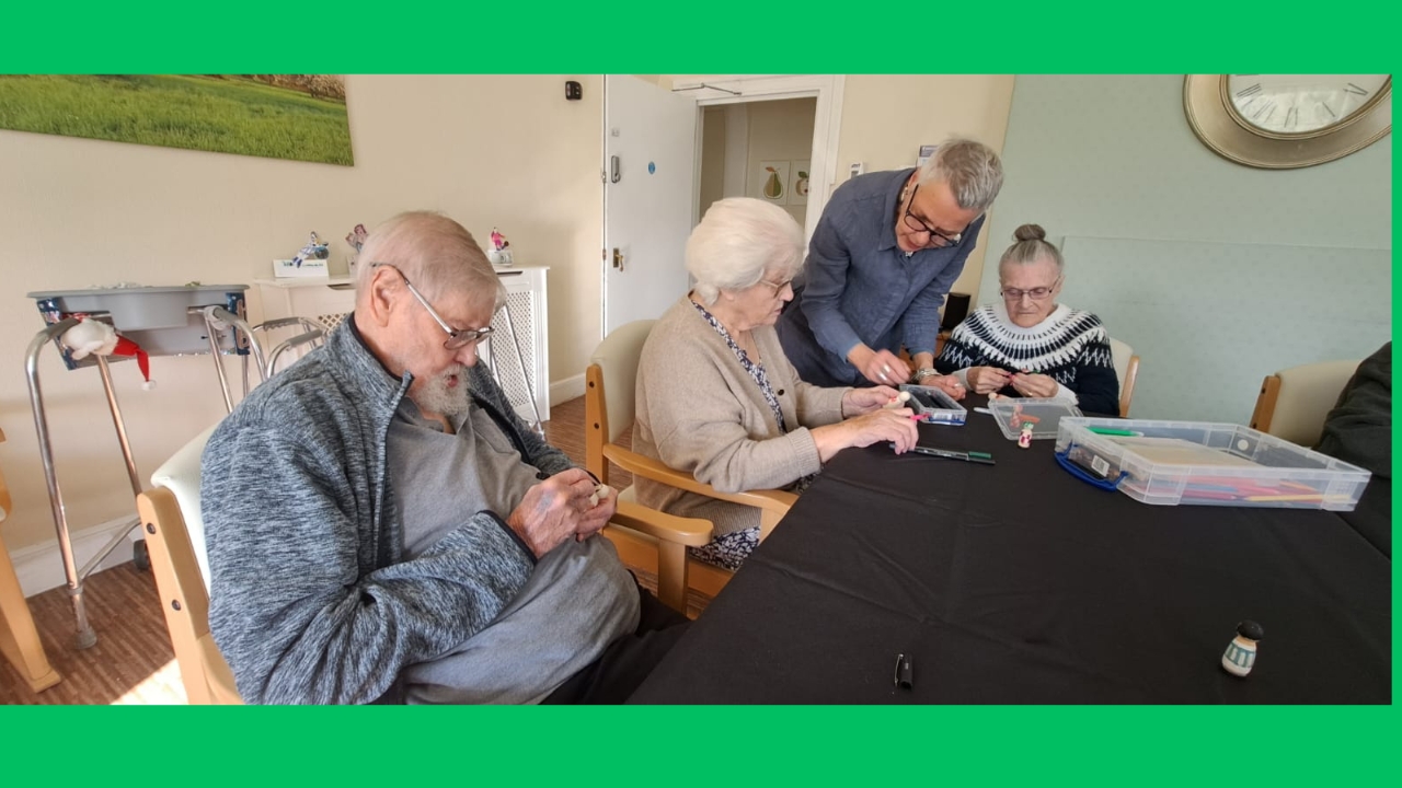 Three people seated at a black-draped table, holding something small in their hands and pens, clearly colouring them in. Each is intent on their work whilst a fourth person stands by offering support