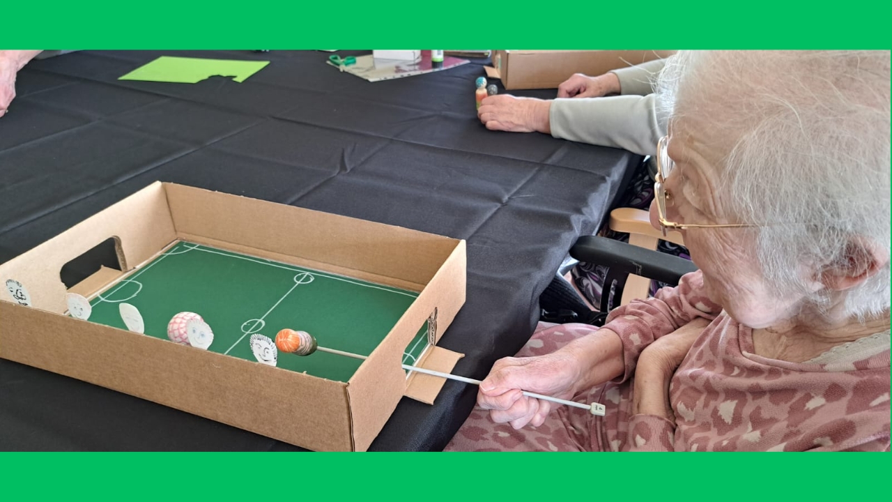 A woman, seated, holding a knitting needle with a figure on the end, inserted into a cardboard football pitch