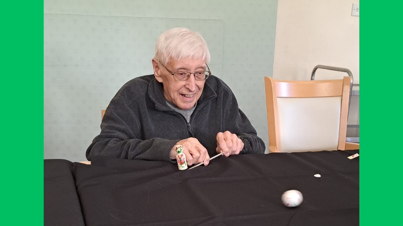 A gentleman seated at a table covered in a black cloth with patterned grey/green wall behind and empty chair beside him. He holds a small wooden figure attached to a knitting needle and is using it to play football across the table using a small white ball