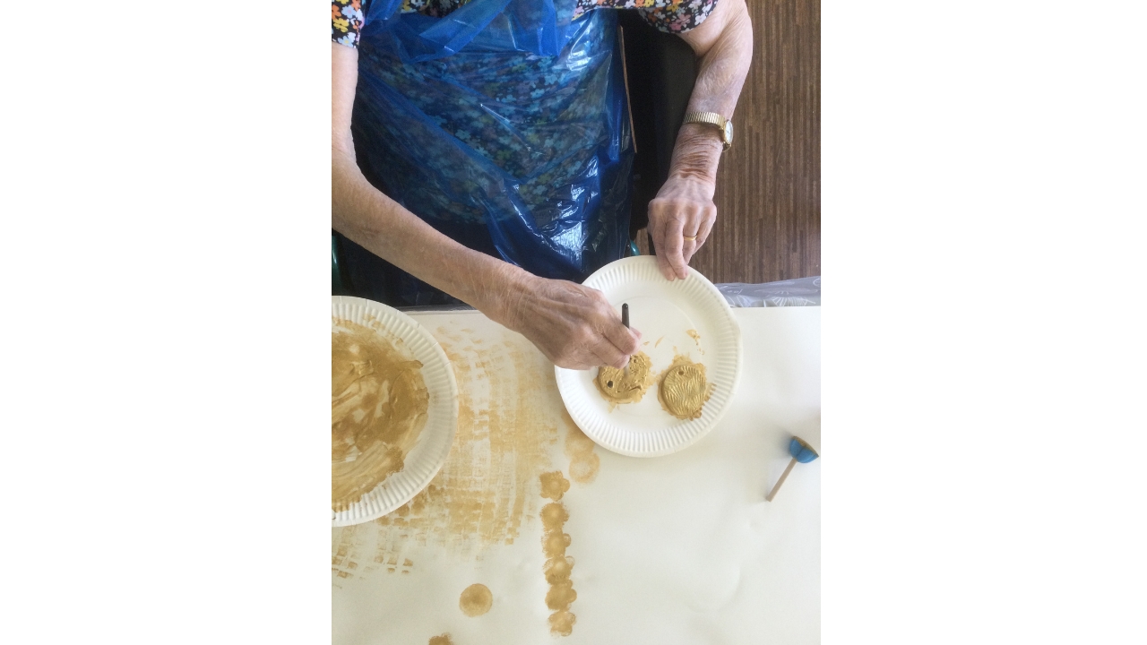 A pair of hands, one holding a paper plate steady whilst the other is using a small paint brush to apply gold paint to two discs on the plate