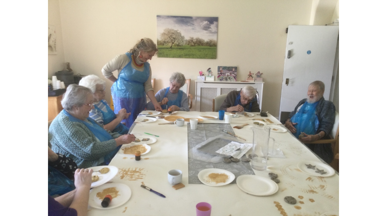 A group of people, most seated, and all wearing blue plastic aprons, around a table with plates of gold paint and assorted brushes