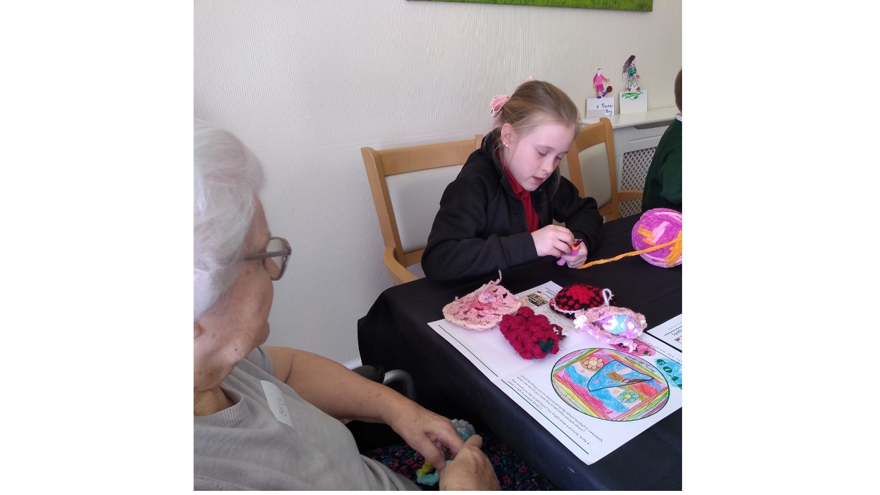 A school girl concentrates intently on a piece of crochet in her hand, a large ball of colourful wool to her let, whilst an adult looks on. In front of her is an array of crocheted items
