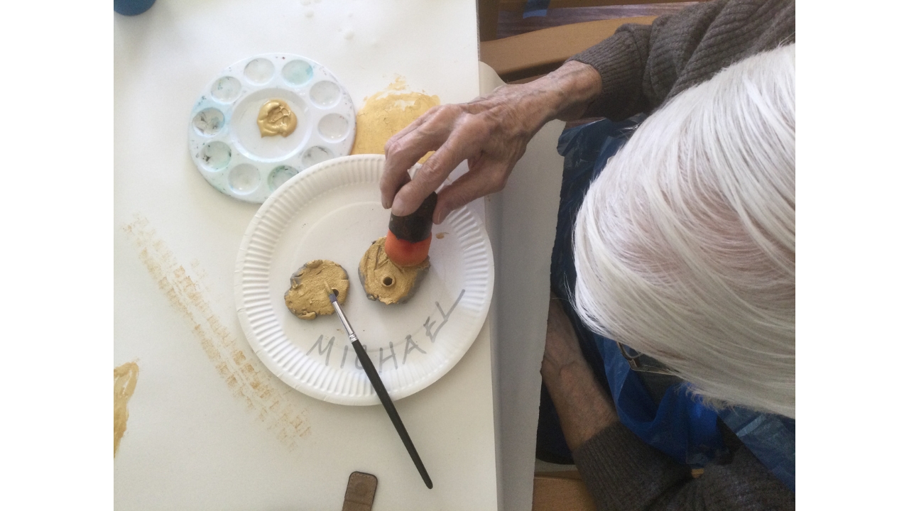 Overhead shot of a whitehaired gentleman with a sponge in his right hand apply gold paint to one of two clay medals on a paper plate in front of him. A small round palette with a smudge of gold paint centre is to his right