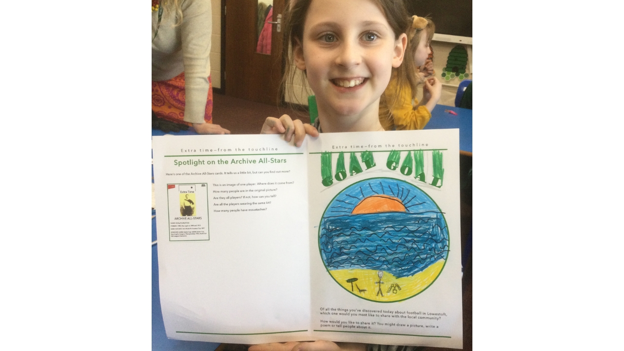 A child holds up her art work and beams at the camera, The picture is a beach scene with sun rising and blue sky and sandy beach